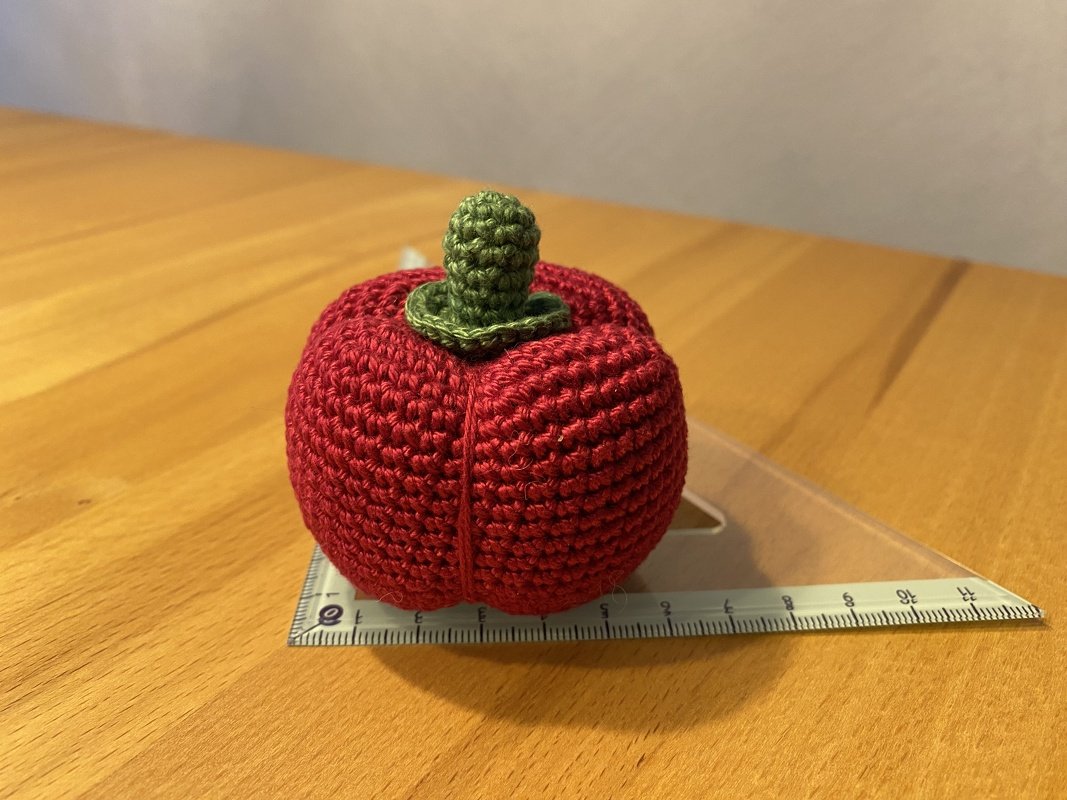 Gehäkelter roter Apfel mit grünem Stiel auf Holztisch, Geodreieck als Größenreferenz.