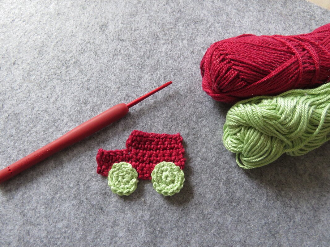 Gehäkelte rote Auto-Applikation mit hellgrünen Rädern, roter Häkelnadel und Garnknäueln auf grauem Filz