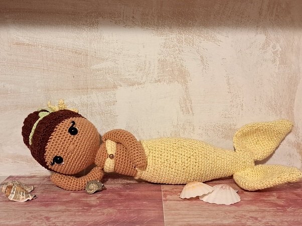 Gehäkelte Amigurumi-Meerjungfrau mit braunen Haaren und hellgelbem Schwanz auf Holz, umgeben von Muscheln.