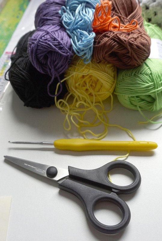 Mehrere bunte Garnknäuel, gelbe Häkelnadel und schwarze Schere auf hellem Untergrund