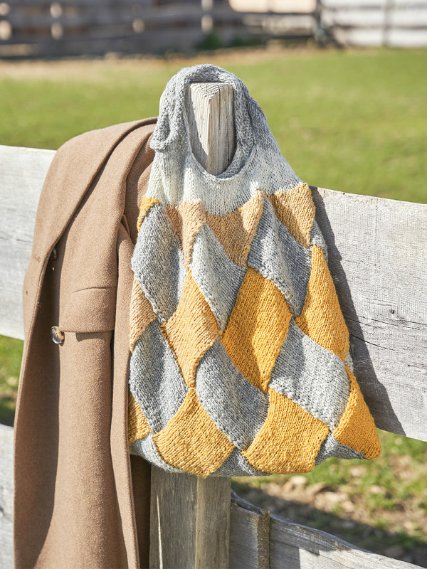 Zweifarbige Stricktasche in Grautönen und Senfgelb mit Rautenmuster, lässig über einen Holzzaun gehängt