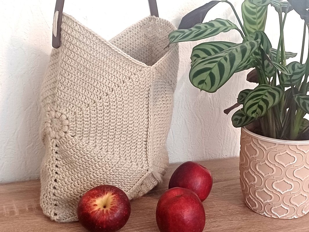 Beigefarbene gehäkelte Umhängetasche mit Blumenmotiv und dunklem Riemen neben roten Äpfeln und einer Topfpflanze
