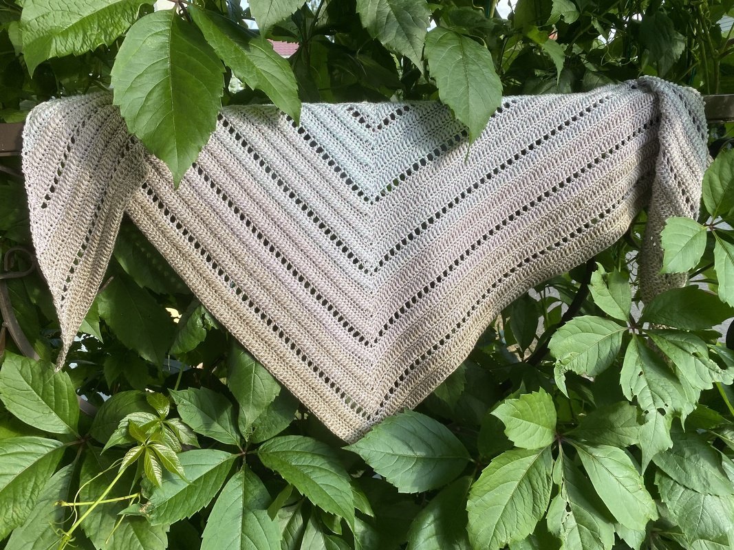 Triangle crochet shawl in neutral variegated yarn with eyelet stripe detail, draped over green leaves