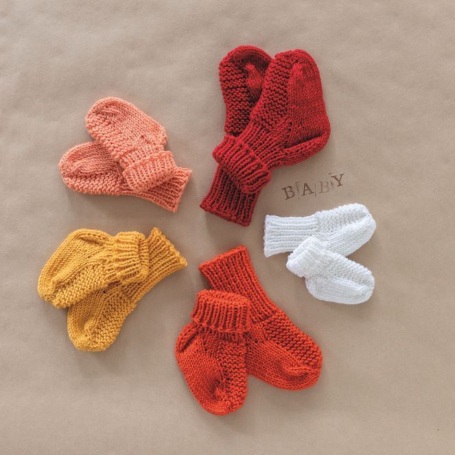 Verschiedene Paare gestrickter Babysocken und Fäustlinge in warmen Farben auf beigem Hintergrund