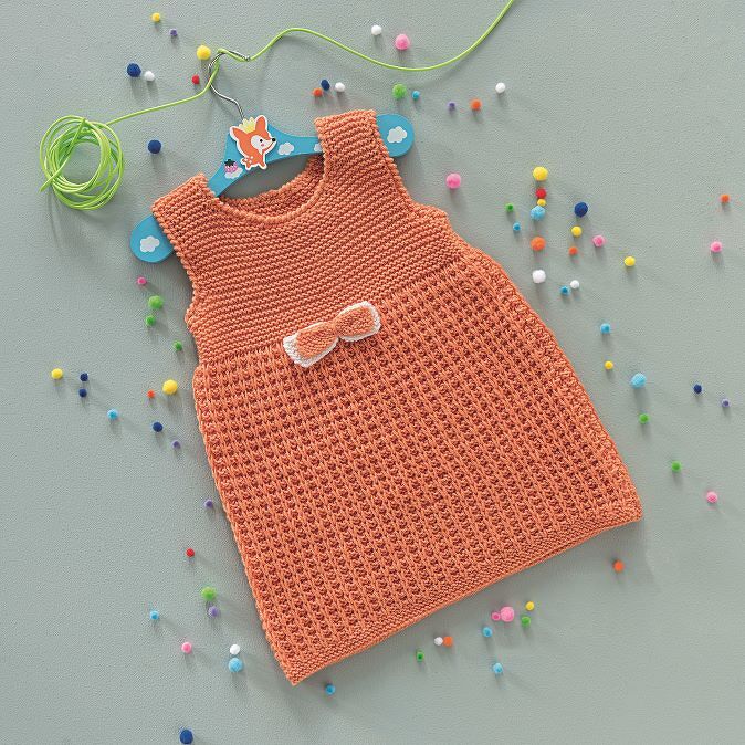 Korallfarbenes gestricktes Babykleid mit Schleife auf blauem Kinderbügel, umgeben von bunten Pompons