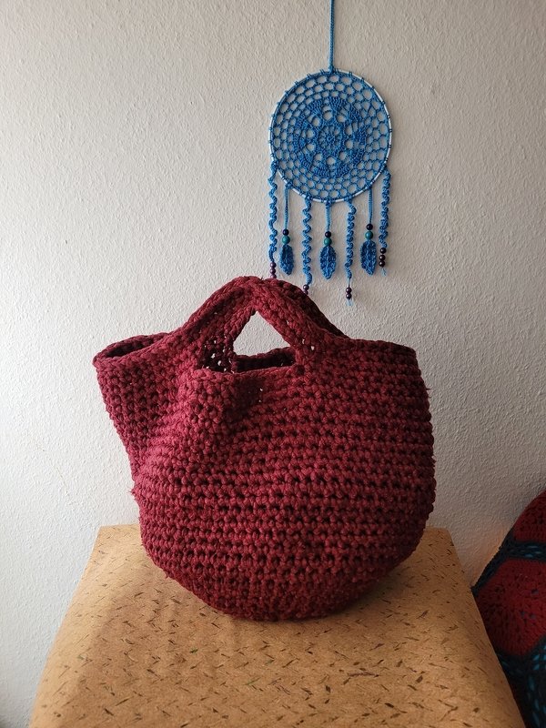 Dunkelrote gehäkelte Shopper-Tasche mit breiten Henkeln auf einer Korkfläche, im Hintergrund ein blauer Traumfänger an einer weißen Wand.