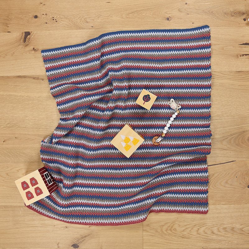 Ansicht einer mehrfarbigen, gestreiften Häkel-Babydecke auf Holzboden mit Holzspielzeug