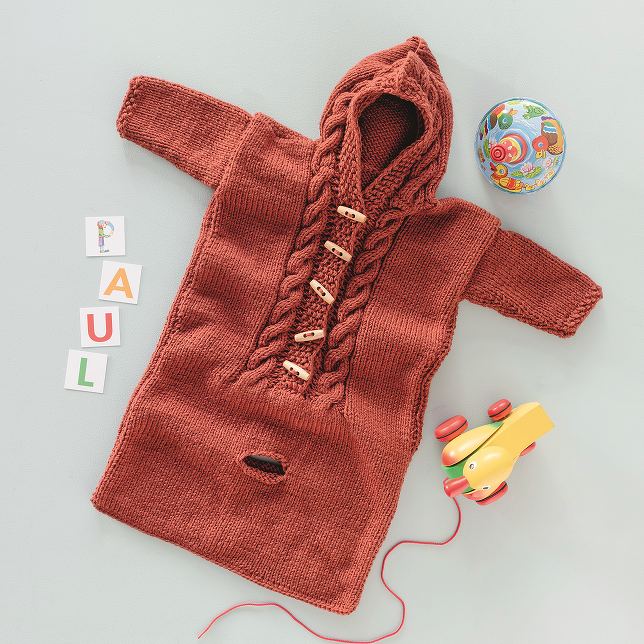 Rostroter gestrickter Babyschlafsack mit Kapuze, Zopfmuster und Holzknöpfen, liegend neben Holzspielzeug