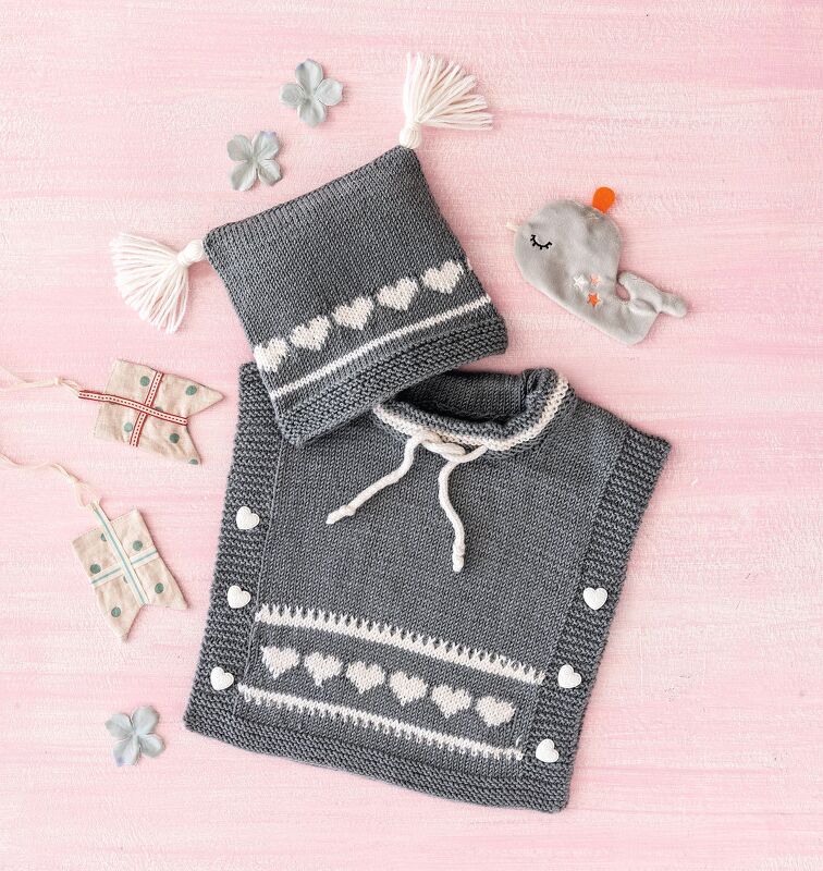 Gestricktes Baby-Set in Dunkelgrau mit weißen Herzen: Mütze mit Quasten und Poncho mit herzförmigen Knöpfen auf rosa Untergrund.