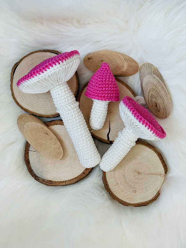 Drei klein gehäkelte Amigurumi‑Pilze mit weißen Stielen und pinken Kappen auf hellen Holzscheiben