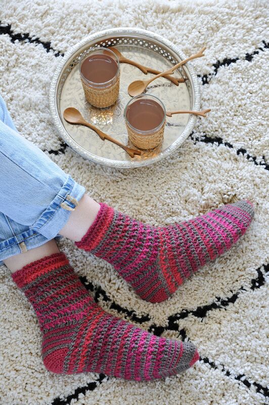 Gehäkelte gestreifte Damensocken in Rosa, Rot und Grau auf einem Teppich neben einem Tablett mit Gläsern.