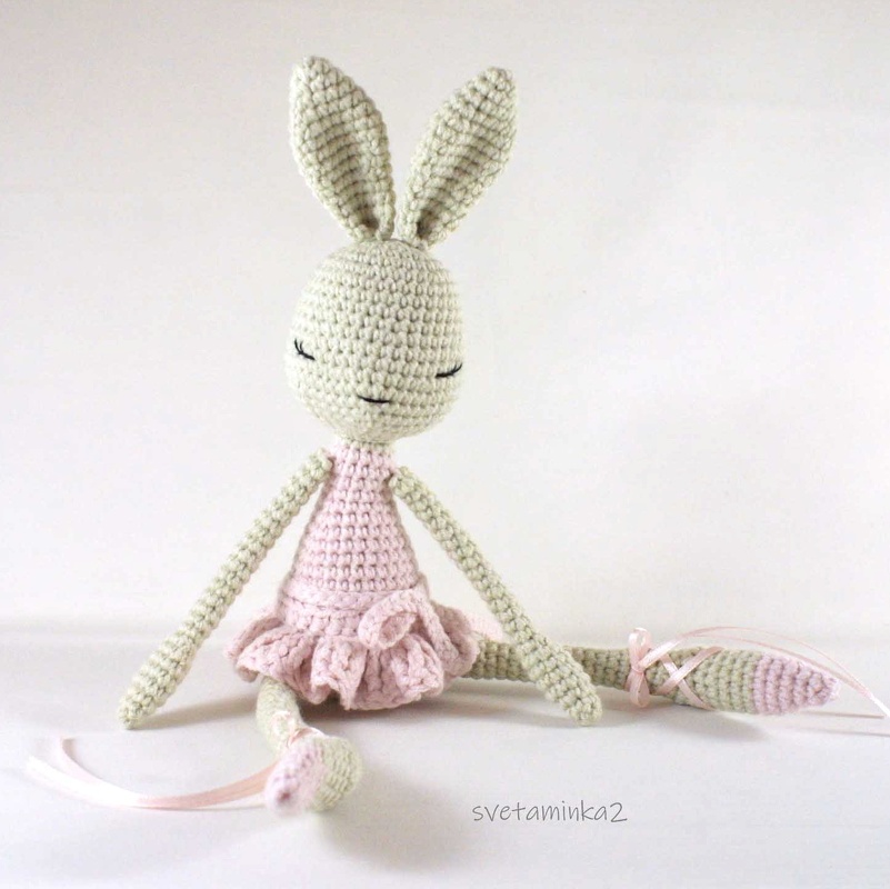 Light green crocheted amigurumi bunny wearing a pink ruffled ballet dress with ribbon-tied ankles, seated on a white background.
