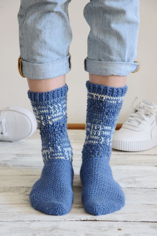 Füße in blauen, gehäkelten Socken mit geometrischem Muster und hochgekrempelten Jeans.