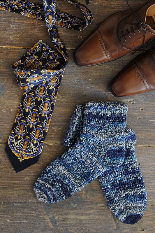 Nahaufnahme handgehäkelter Socken in Blau-Grau-Weiß neben gemusterter Krawatte und braunen Schuhen auf Holz.