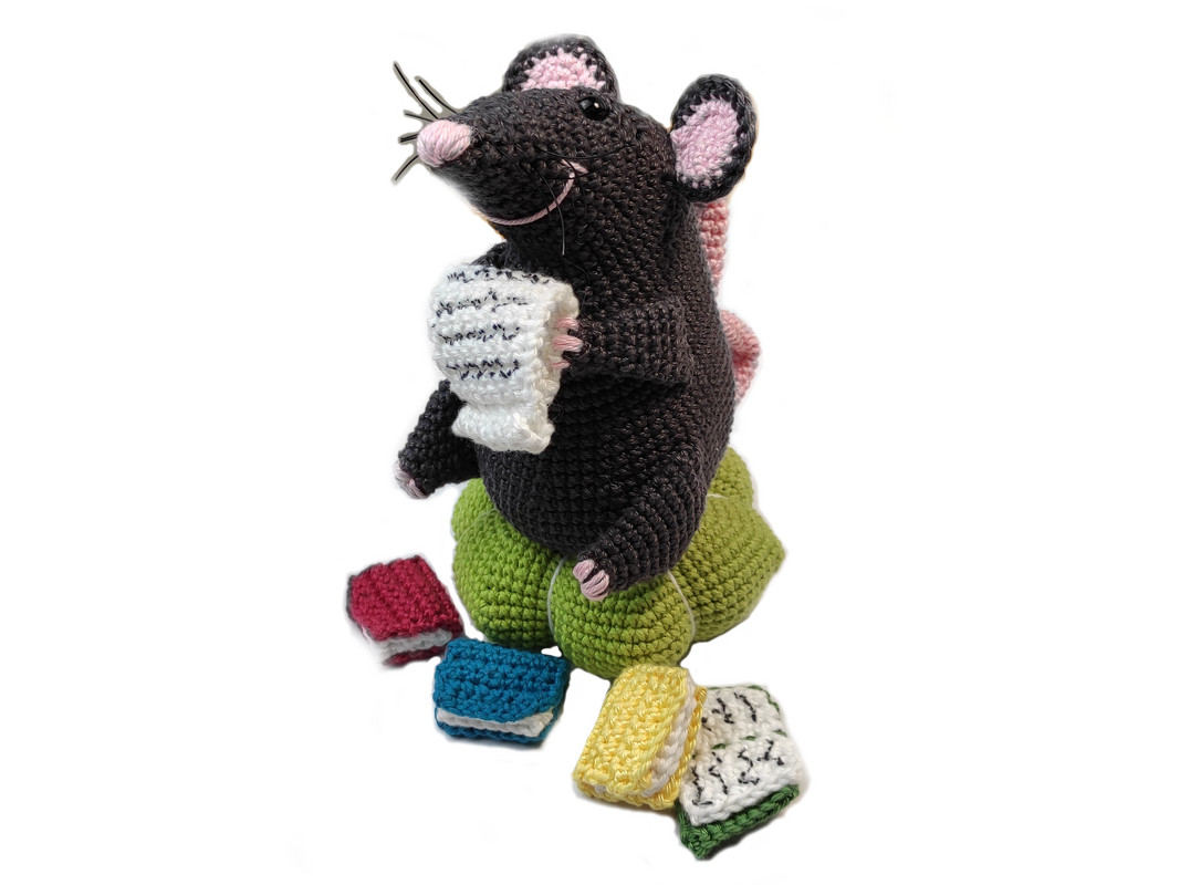 Gehäkelte dunkelgraue Maus mit rosa Ohren auf grünem Kissen, hält weißes gehäkeltes Blatt; bunte Mini‑Bücher davor