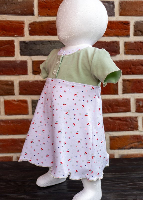 Kurzärmeliges Babykleid mit pastellgrünem Oberteil und weißem Rock mit kleinem Kirschmuster, zweigeteilte Knopfleiste vorne, auf Schaufensterpuppe vor Ziegelwand