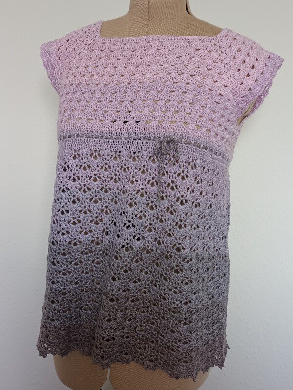 Gehäkelte Damen-Tunika in Flieder zu Grau auf Schneiderpuppe, mit eckigem Ausschnitt, kurzen Ärmeln und Spitzenrand am Saum.