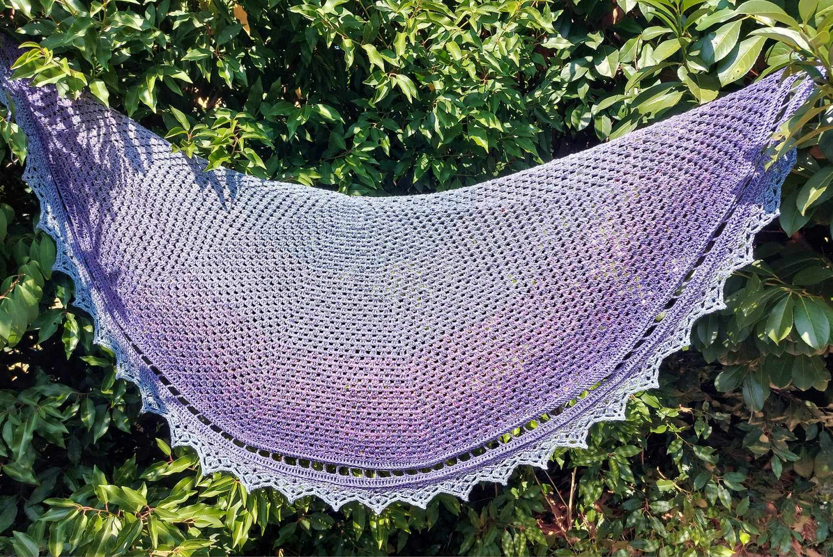 Halbrundes, gehäkeltes Tuch mit Lila‑Blauem Farbverlauf und gezackter Borte, vor grünen Blättern aufgehängt.