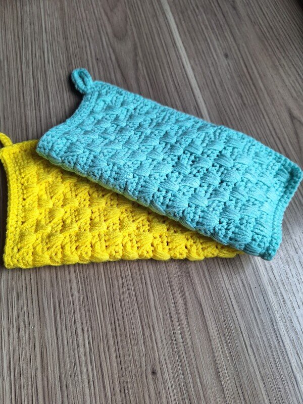 Zwei gehäkelte Topflappen, gelb und türkis, mit Strukturmuster und Aufhängeschlaufen auf Holzuntergrund.