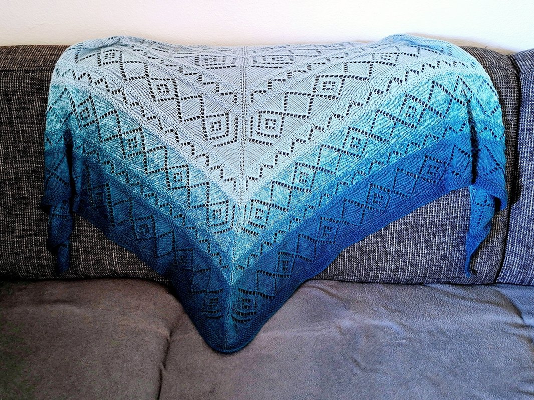 Dreieckstuch gestrickt mit Ajour‑Rautenmuster, Farbverlauf von hell- zu dunkelblau, auf grauem Sofa drapiert