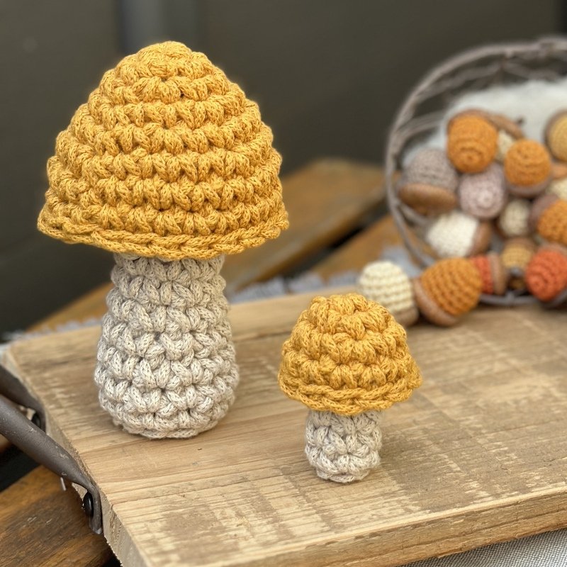 Zwei gehäkelte Pilze mit senfgelben Kappen und hellbeigen Stielen auf einem Holzbrett