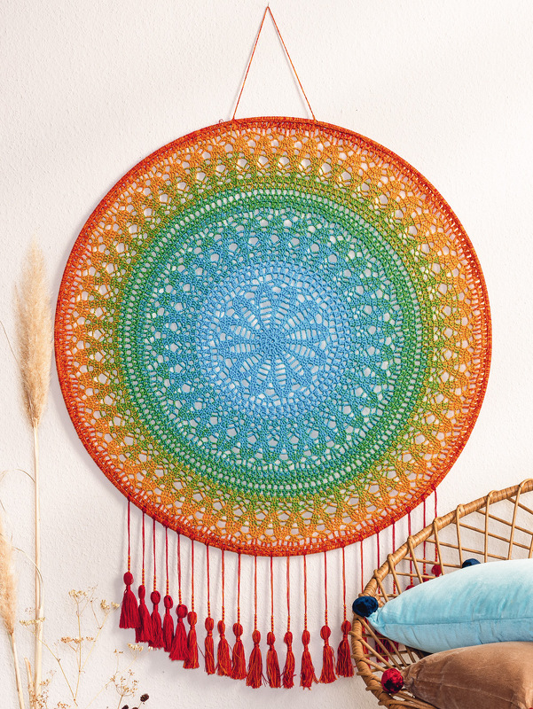 Rundes gehäkeltes Mandala mit Farbverlauf und roten Quasten an weißer Wand