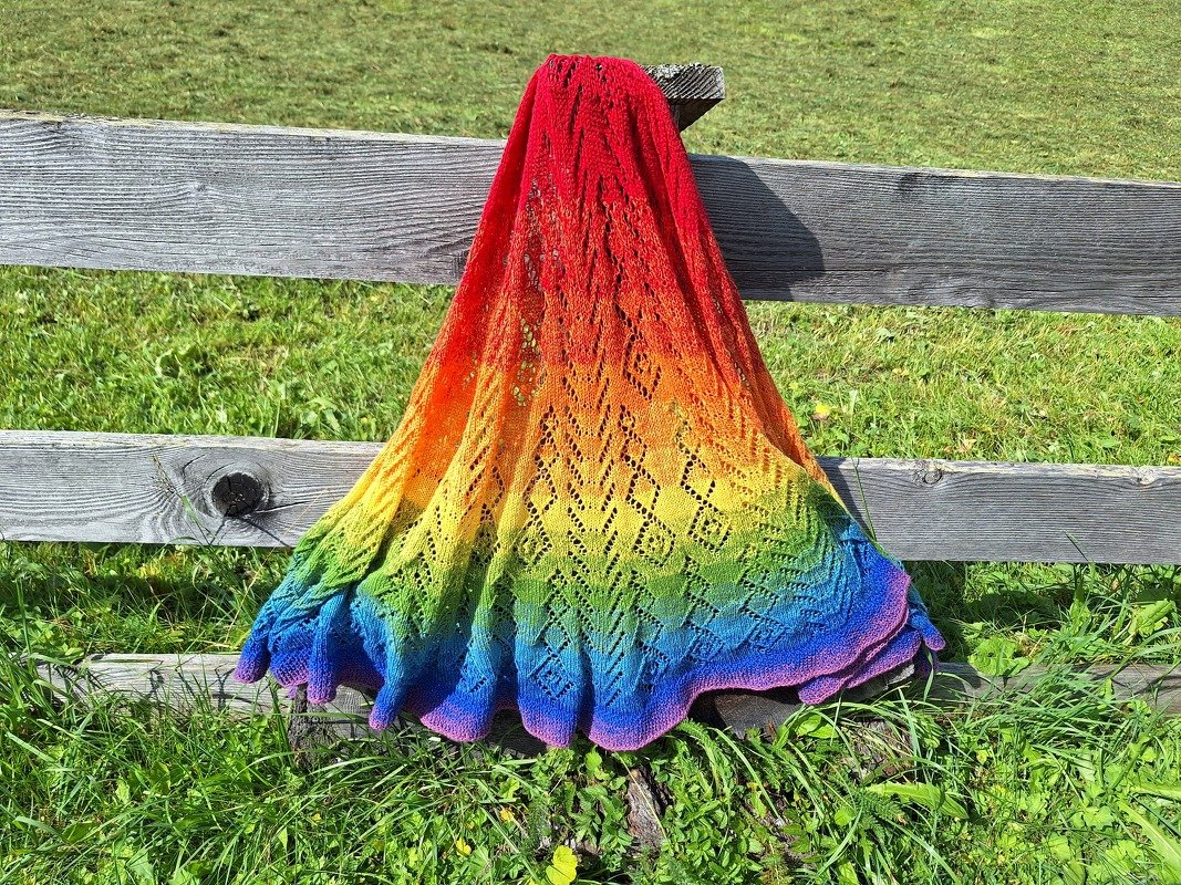 Gestricktes Lacetuch in Regenbogenfarben mit Ajour-Lochmuster, über einem Holzzaun drapiert, Gras im Hintergrund