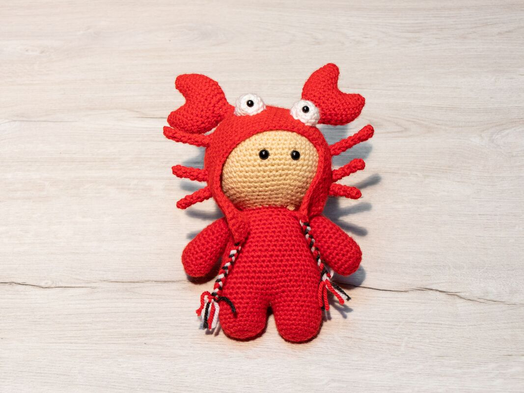 Gehäkelte Amigurumi-Krabbe in rotem Kostüm auf hellem Holzhintergrund.