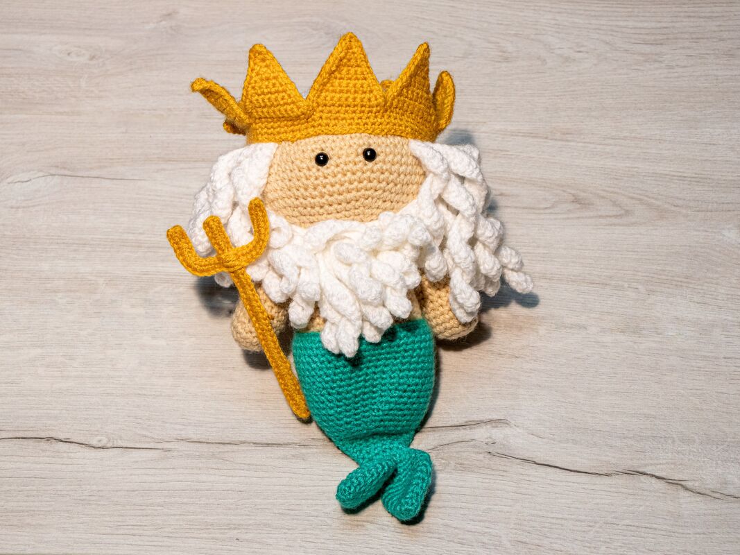 Gehäkelter Amigurumi-Meereskönig mit goldgelber Krone, weißem Bart, türkisgrüner Flosse und Dreizack.