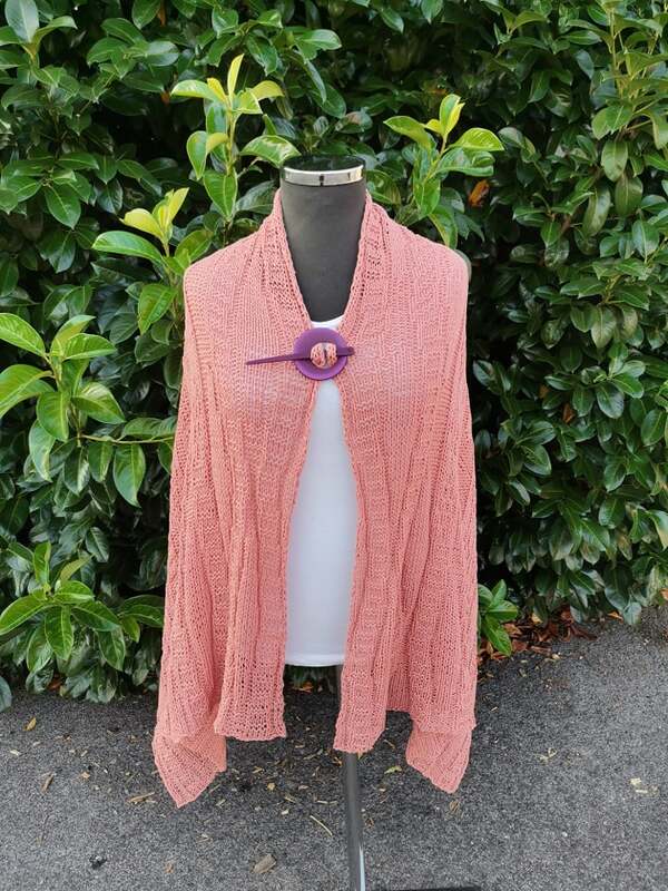 Korallfarbenes gestricktes Umschlagtuch mit Lochmuster, befestigt mit violetter Brosche vor grünem Laub