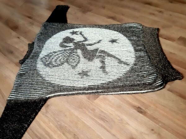 Damen-Strickjacke mit großem Intarsienkreis und Silhouette einer fliegenden Figur auf einem Besen, dunkle und helle Töne, flach auf Holzboden gelegt.