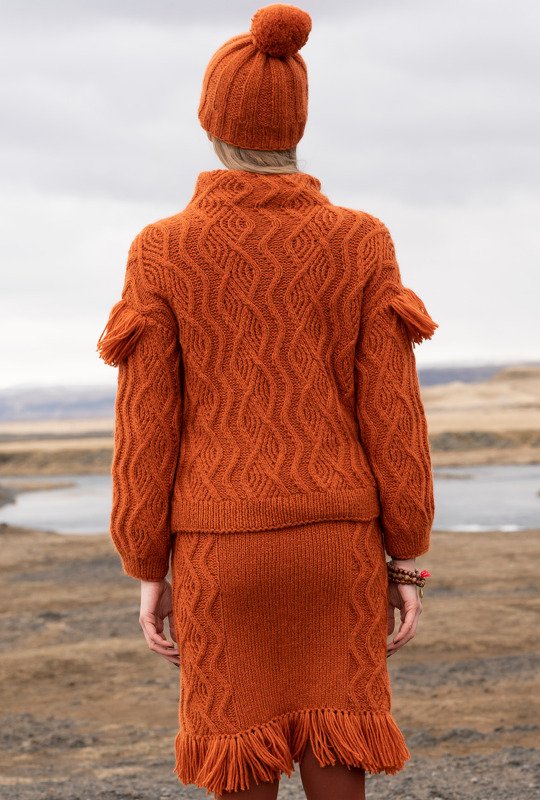 Rückenansicht: Frau in rostorangefarbenem Strickpullover, Rock mit Fransen und Bommelmütze vor einer Naturlandschaft.