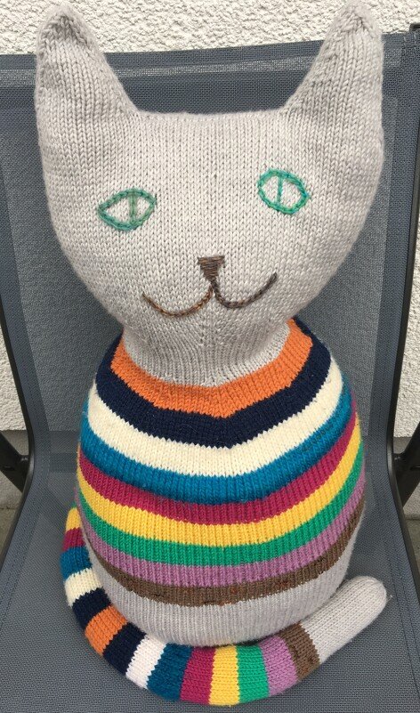 Gestrickte Amigurumi-Katze mit hellgrauem Kopf und bunten, horizontalen Streifen am Körper und Schwanz, sitzt auf einem Stuhl.
