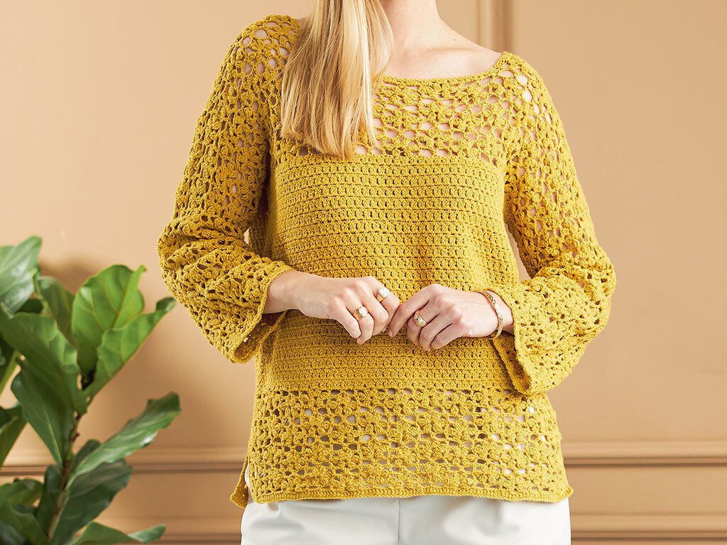 Frau in senfgelbem gehäkeltem Pullover mit Lochmuster und hellen Hosen vor beiger Wand