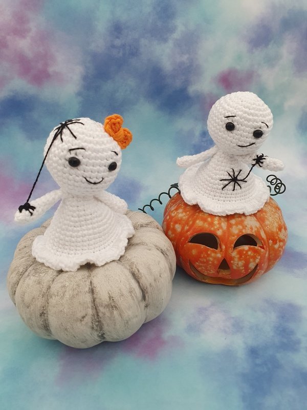 Two white crocheted ghost amigurumi with stitched faces and an orange bow sitting on decorative pumpkins against a pastel backdrop