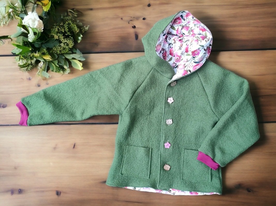Grüne, gefütterte Kinderkapuzenjacke mit floralem Innenfutter, aufgesetzten Taschen und magentafarbenen Bündchen, liegend auf Holzuntergrund
