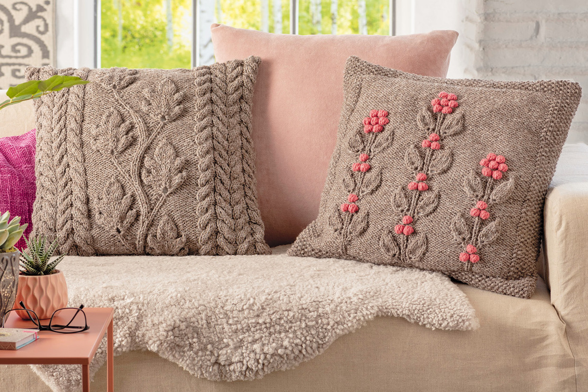 Gestrickte Kissen mit Zopf- und Blattmuster sowie rosa Blüten auf Sofa