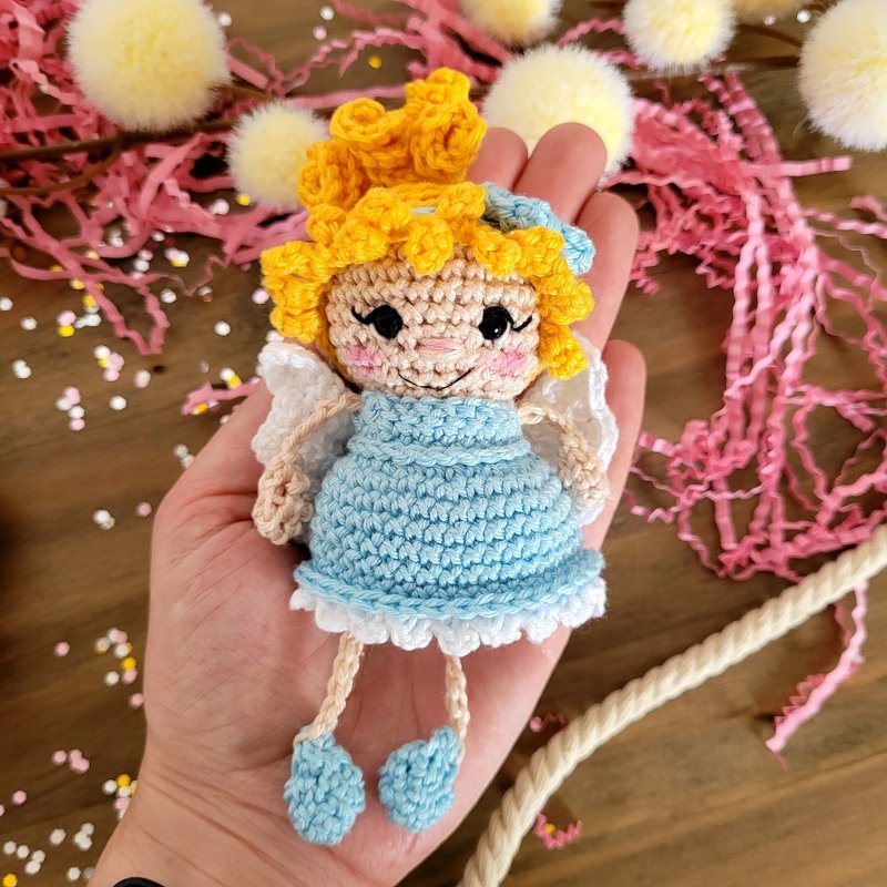 Hand hält kleines gehäkeltes Engel-Amigurumi mit gelben Locken, blauem Kleid und weißen Flügeln