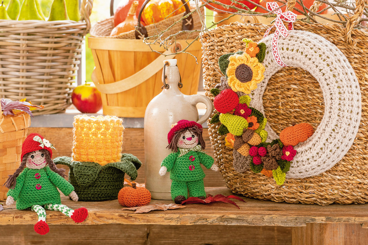 Herbstliche Häkeldeko mit zwei Amigurumi-Wichteln, kleinem gehäkelten Kürbis und Herbstkranz mit Sonnenblume und Fruchtmotiven auf einem Holztisch.