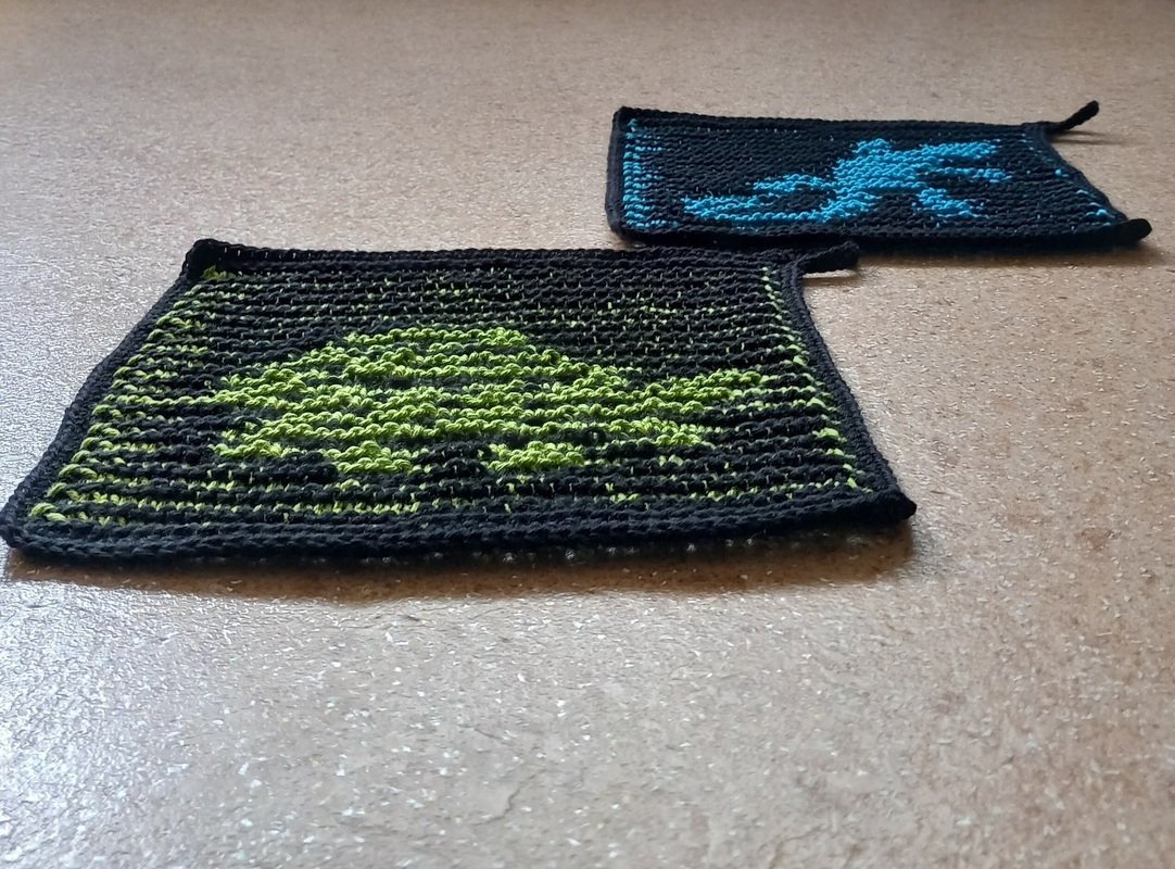 Zwei gestrickte Topflappen auf hellem Untergrund: einer mit grünem Baum- bzw. Drachenmotiv auf dunklem Grund, der andere mit blauem Krabben- bzw. Hummermotiv.