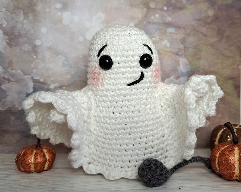 Weißes gehäkeltes Amigurumi-Gespenst mit schwarzen Augen, rosa Wangen und gewelltem Saum neben Dekokürbissen