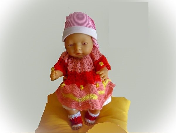 Babypuppe auf gelbem Kissen trägt gehäkeltes rosa-rotes Kleid mit gelben Wellenstreifen, gestreifte Mütze und kleine Söckchen