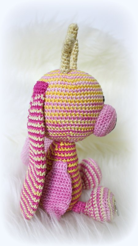 Gehäkeltes Amigurumi-Tier, pink und gelb gestreift, sitzend auf weißem, flauschigem Untergrund