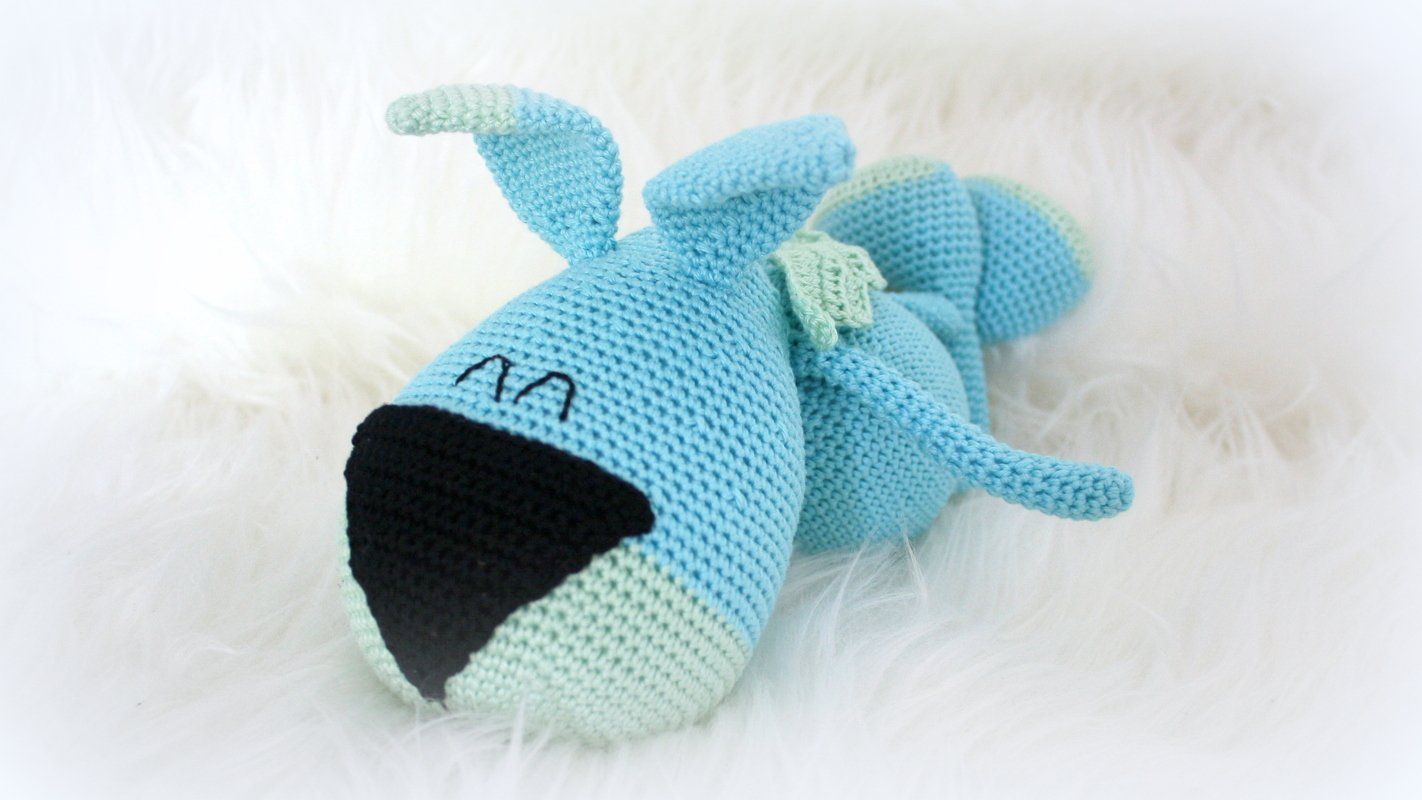 Gehäkelter hellblauer Amigurumi-Hund liegend mit schwarzer Nase und gestickten Augen