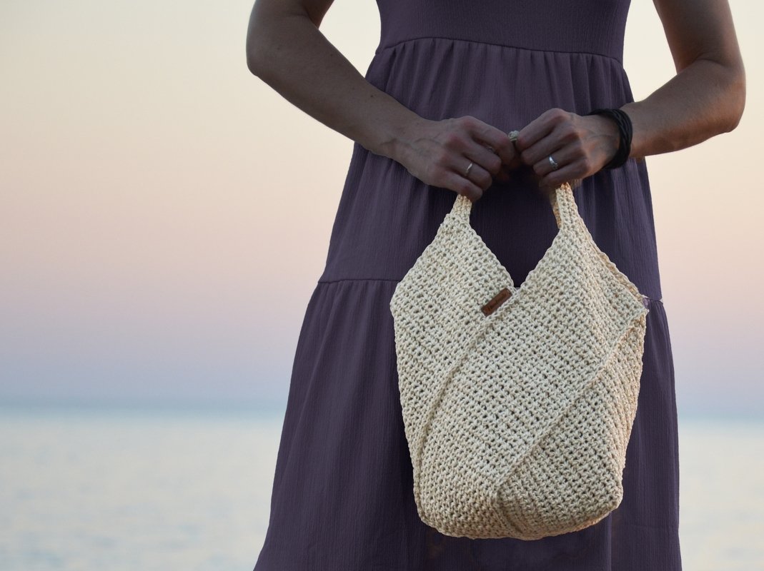 Handgehäkelte, cremefarbene Shopper-Tasche mit strukturierter Oberfläche, gehalten vor dem Meer bei Sonnenuntergang.
