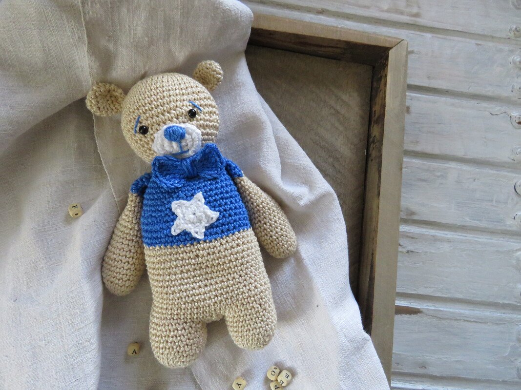 Gehäkelter beiger Amigurumi-Teddybär mit blauem Pullover und weißem Stern, liegend auf Stoff, zeigt Gesamtproportionen.