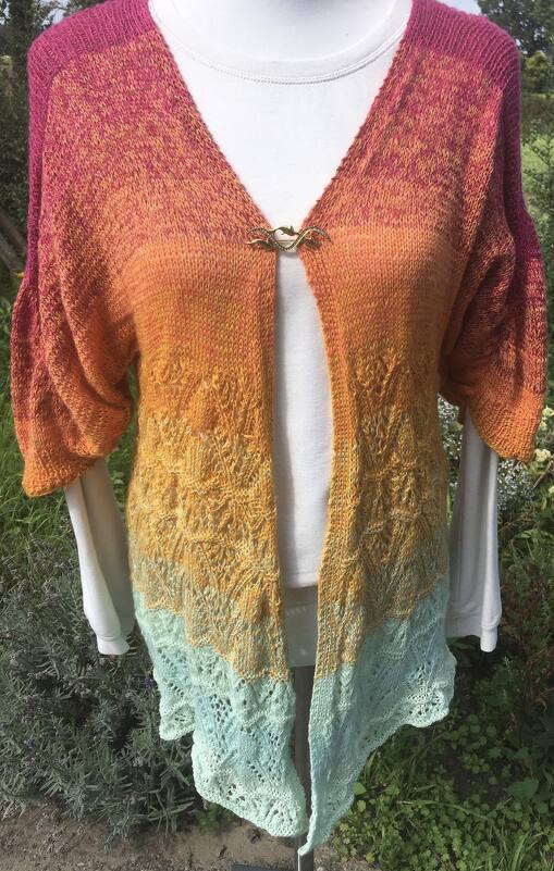 Offene gestrickte Damenjacke mit Farbverlauf von Pink zu Türkis, dekorativer Spange und filigranem Spitzenmuster am Saum.