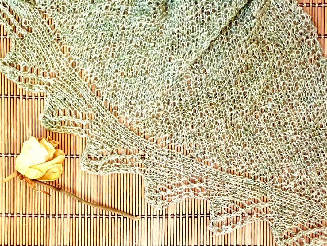 Gestricktes Dreieckstuch mit lockerem Lochmuster und gezackter Bordüre, liegend auf einer Bambusmatte neben einer getrockneten gelben Rose