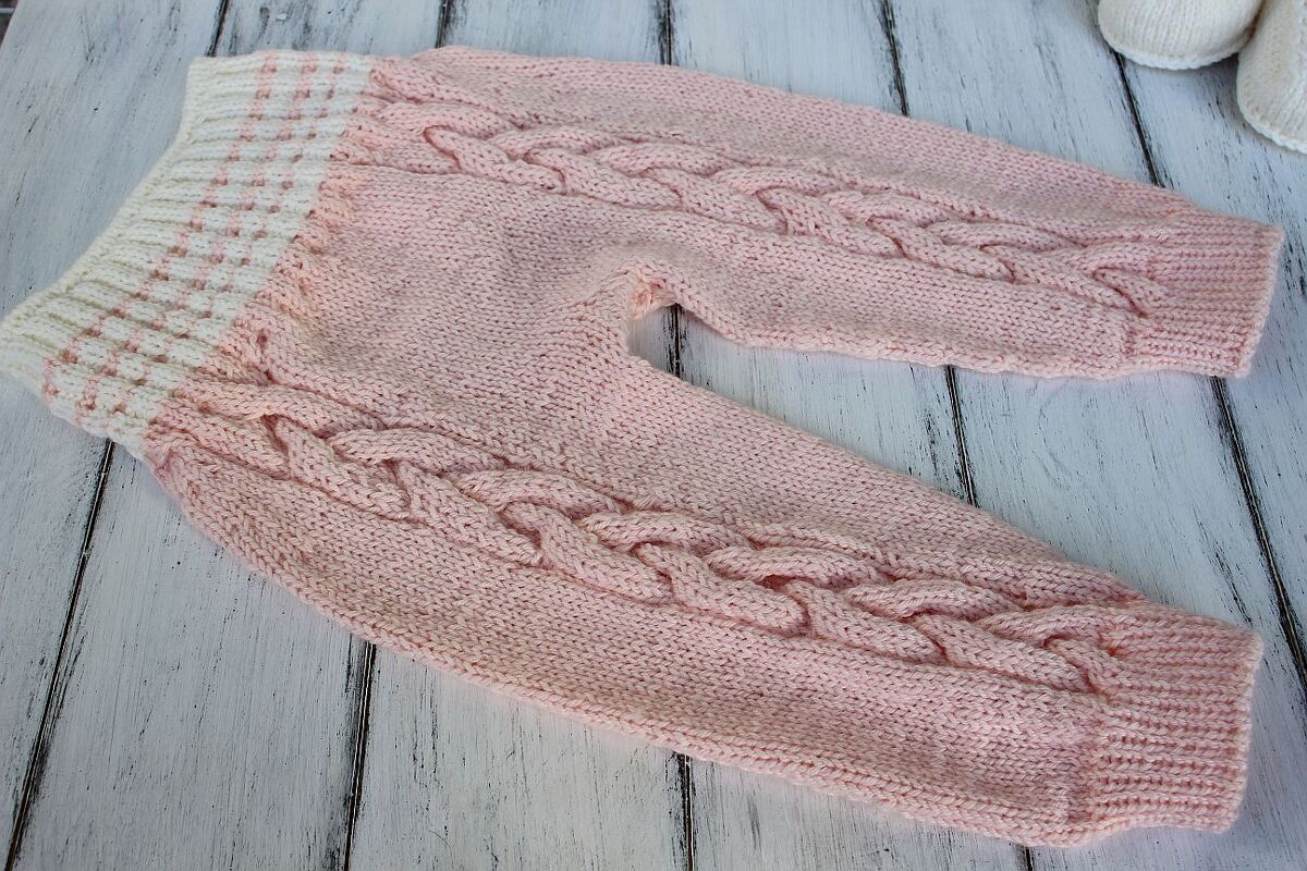 rosa gestrickte Babyhose mit Zopfmuster und weiß-rosa Bündchen auf hellem Holz