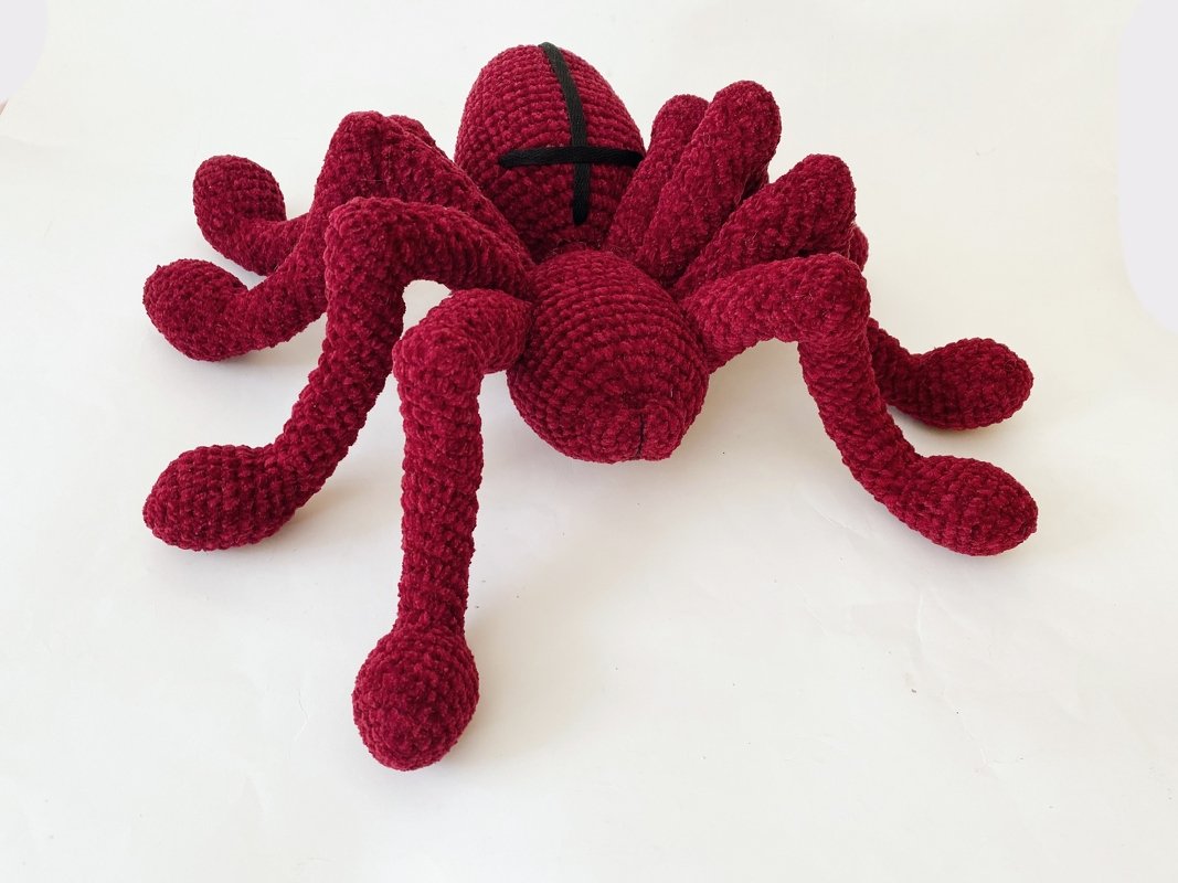 Weinrote gehäkelte Amigurumi-Spinne mit acht dicken Beinen und schwarzem Kreuzmuster auf hellem Hintergrund.